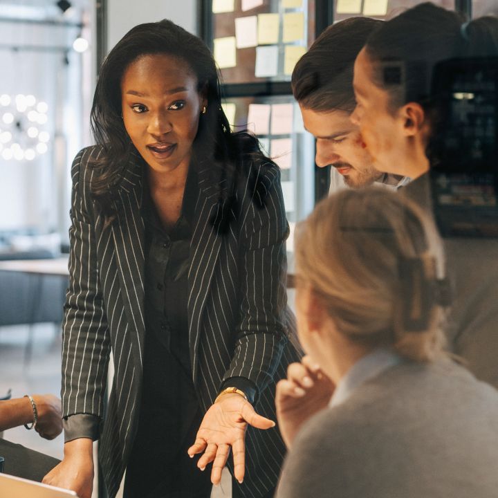 Group of professionals gathered around a table in a meeting room, discussing ideas in front of a glass wall covered with sticky notes.