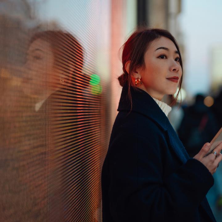 Confident young Asian woman with smartphone, standing against digital display on the street in the city at night.